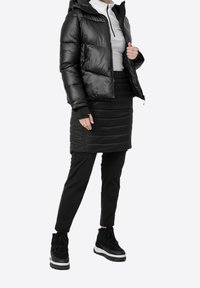 Schwarze Daunenjacke mit glänzendem Finish, taillierte Silhouette, Reißverschluss vorne, kombiniert mit einem schwarzen Rock und einer Hose, getragen mit schwarzen Sneakers.