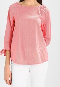 Blouse rayée rouge et blanche avec un décolleté bateau, des manches trois-quarts et des détails de lien au niveau des poignets. Tissu léger avec une texture douce.