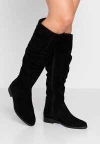 Czarne zamszowe kozaki do kolan o luźnym kroju, z płaską podeszwą i suwakiem z boku. Teksturowana powierzchnia z gładkim akcentem na palcach.