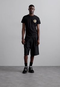 Μαύρο μπλουζάκι κοντό μανίκι με στρογγυλό χρυσό λογότυπο στο στήθος, συνδυασμένο με φαρδιά μαύρα σορτς και μαύρα αθλητικά παπούτσια.