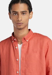 Camicia leggera in lino corallo con colletto button-down, dotata di bottoni bianchi e un logo sottile sul lato sinistro del petto. Indossata sopra una t-shirt bianca.