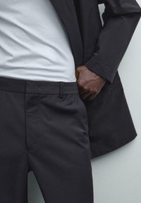 Man in zwarte broek en zwart colbert met een wit shirt in zijn broek, hand op de heup tegen een lichte achtergrond.
