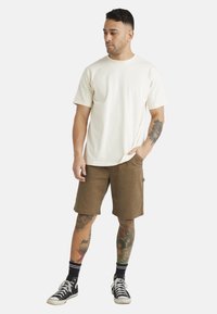 Kortærmet creme t-shirt parret med brune shorts. Outfittet har en afslappet pasform, med sneakers og synlige farverige tatoveringer på benene.