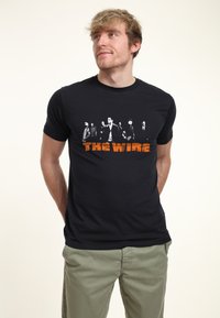 Svart bomullst-shirt med en grafisk tryck av karaktärer från "The Wire" i svart och vitt med orange tryck. Kort ärm, rund hals.