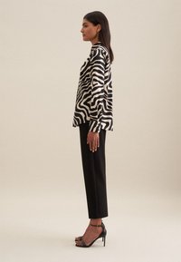 Blusa di seta a righe bianche e nere con colletto, indossata con pantaloni neri su misura e sandali neri con tacco alto. Vista di profilo.