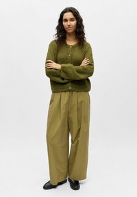 Vihreä neulottu neuletakki, jossa etupuolella napit, yhdistetty vaaleisiin khakihousuihin, joissa on laskokset. Asusteina mustat loafersit. Yksinkertainen, teksturoitu muotoilu.