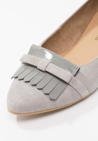 Zapato plano de ante gris claro con puntera afilada, que presenta un fleco gris brillante y un detalle de lazo de ante a juego.