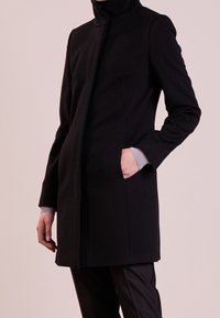 Sort uldfrakke med høj krave, lange ærmer og sidelommer. Glat tekstur og tætsiddende design med minimal detaljering.