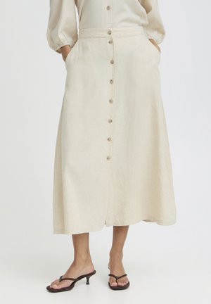 Naine kannab beežikat keskpikkuse ees nööbitava seelikuga, käed taskutes ja pruunides kontsakingades.