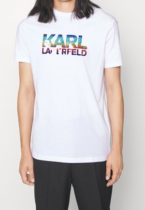 Persoon draagt een wit T-shirt met een meerkleurig "KARL LAGERFELD" logo, gecombineerd met een zwarte broek tegen een effen achtergrond.