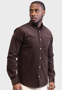 Camisa de pana marrón con cuello abotonado, mangas largas, bolsillo frontal y botones blancos. Textura suave, corte entallado.