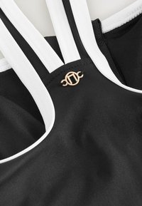 Mustkollariga sporditopp valgete triipudega, millel on struktureeritud kangas. Kuldsest logost aktsent asub rihmade ristumiskoha lähedal.