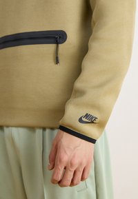 Μπεζ φούτερ με μαύρη τσέπη με φερμουάρ, ελαστικούς καρπούς και λογότυπο Nike στο μανίκι. Μαλακό ύφασμα και άνετη εφαρμογή. Φοριέται με ανοιχτό πράσινο παντελόνι.