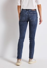 Jean en denim bleu skinny avec une taille mi-haute, des poches arrière et une texture de lavage discrète, associés à des sneakers blanches et beiges.