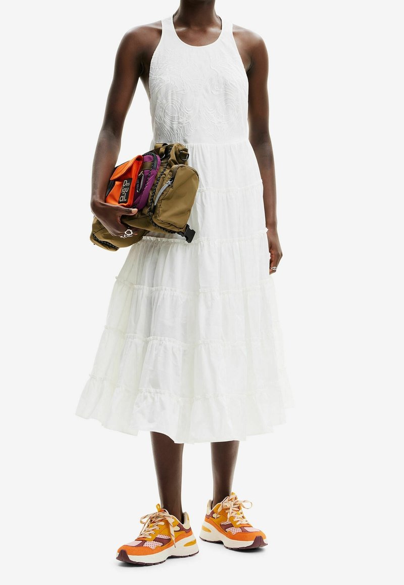 Vestido maxi blanco con capas y textura bordada, combinado con zapatillas naranjas y crema. La modelo sostiene una mochila marrón y colorida.