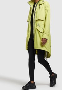 Ljusgul vattentät jacka med dragkedja fram, elastiska ärmslut och huva, i kombination med svarta leggings och svarta sneakers.