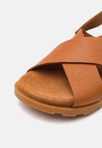 Närbild av en brun lädersandal med korsade remmar och en vadderad sula på en vit bakgrund.