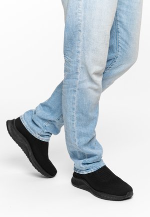 Person iført lyseblå jeans og sorte slip-on strikkede sneakers, der går på en hvid baggrund.