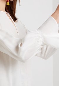 Vit långärmad blus med samlade manschetter och detalj med nyckelhål vid halsen, tillverkad av ett lätt, genomskinligt tyg.