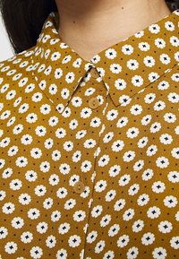 Camicia con colletto in tessuto giallo senape, decorata con piccoli modelli a fiori bianchi con accenti neri e bottoni marroni lungo la parte anteriore.
