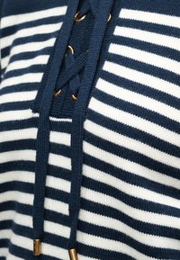 Pull tricoté rayé bleu marine et blanc avec un détail avant à lacets bleu marine entouré d'œillets dorés et d’embouts métalliques de cordon.