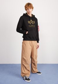 Felpa nera con logo dorato, pantaloni beige a gamba larga e sneakers neri/bianchi. Texture liscia e vestibilità rilassata con dettagli di cucitura visibili.