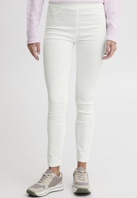 Bele skinny jeans z gladko teksturo, ki imajo sprednje žepe in visok pas, v kombinaciji s svetlo sivo obutvijo.