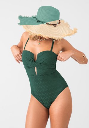 Maillot de bain une pièce vert foncé avec un motif zigzag texturé, doté d'un décolleté en cœur et d'un détail en trou de serrure, associé à un chapeau en paille à large bord.