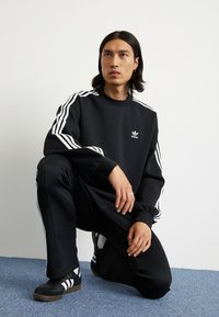 Svart sweatshirt med vita ränder på ärmarna, kombinerad med svarta byxor och svarta sneakers med vita detaljer. Adidas-logotypen framtill.