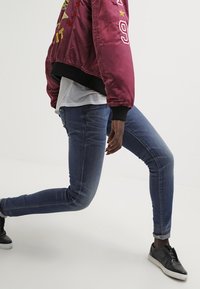 Burgundské saténové bomber bundy s výšivkou, kombinované s přiléhavými modrými džíny a černými teniskami. Džíny mají mírně potrhaný vzhled.