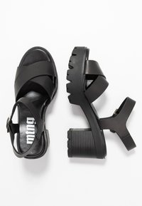 Sandales noires à lanières avec un talon épais, en matériau synthétique lisse, avec des lanières croisées et une plateforme texturée pour une meilleure adhérence.