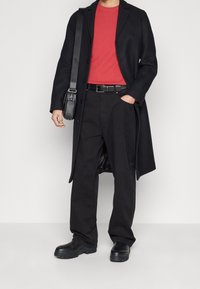 Svart lång kappa över en röd skjorta, kombinerat med svarta byxor och kraftiga svarta stövlar. En axelväska fulländar outfiten.