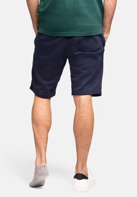 Pantaloni corti blu navy con vestibilità rilassata, realizzati in tessuto morbido, dotati di tasche laterali e dettaglio della tasca posteriore. Abbinati a calzini grigi e scarpe bianche.