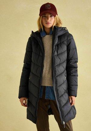 Čierna puffer bunda s prešívaným vzorom, s uzáverom na zips a kapucňou. V kombinácii s béžovým svetrom a džínsovou košeľou, nosená s kockovanými nohavicami. Bordová čiapka.