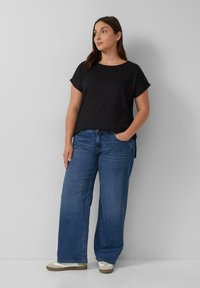 Zwart gestructureerd T-shirt met korte mouwen, gecombineerd met losse blauwe jeans en beige sneakers, weergegeven tegen een neutrale achtergrond.
