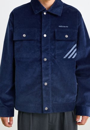 Granatowa kurtka sztruksowa z przednimi zatrzaskami, dwoma kieszeniami na piersi, logo Adidas oraz trzema jasnoniebieskimi ukośnymi paskami po prawej stronie.