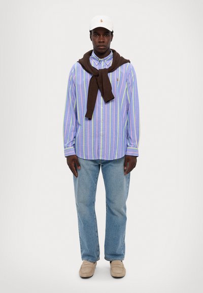 Chemise rayée à boutons de couleur bleu clair, avec des rayures vertes et violettes discrètes. Pull marron drapé sur les épaules, pantalon en denim bleu clair, chaussures beiges.
