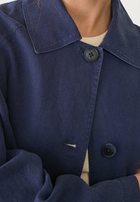 Marineblauwe, geweven jas met een grote kraag, twee knopen en een zijsplit. De stof lijkt gestructureerd en licht gekreukt.