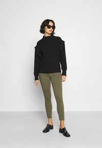 Sweater preto canelado com gola alta, calças justas verde-oliva e mules pretos de salto. Os acessórios incluem óculos de sol escuros.