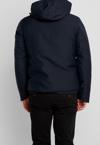 Uomo in giacca con cappuccio blu navy e pantaloni neri, di spalle, in piedi contro uno sfondo grigio chiaro uniforme.