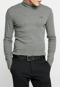 Maglione grigio a collo alto realizzato in tessuto morbido, con maniche lunghe e una vestibilità aderente. Abbinato a pantaloni neri e una cintura nera.