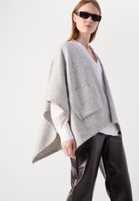 Lysegrå poncho lavet af blød uld, med en frontlomme og en afslappet pasform, parret med en hvid bluse og skinnende sorte bukser.