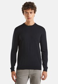 Černý pletený svetr s dlouhými rukávy a kulatým výstřihem. Má hladkou texturu a žebrované manžety a spodní lem. Nosený přes světlé kalhoty.