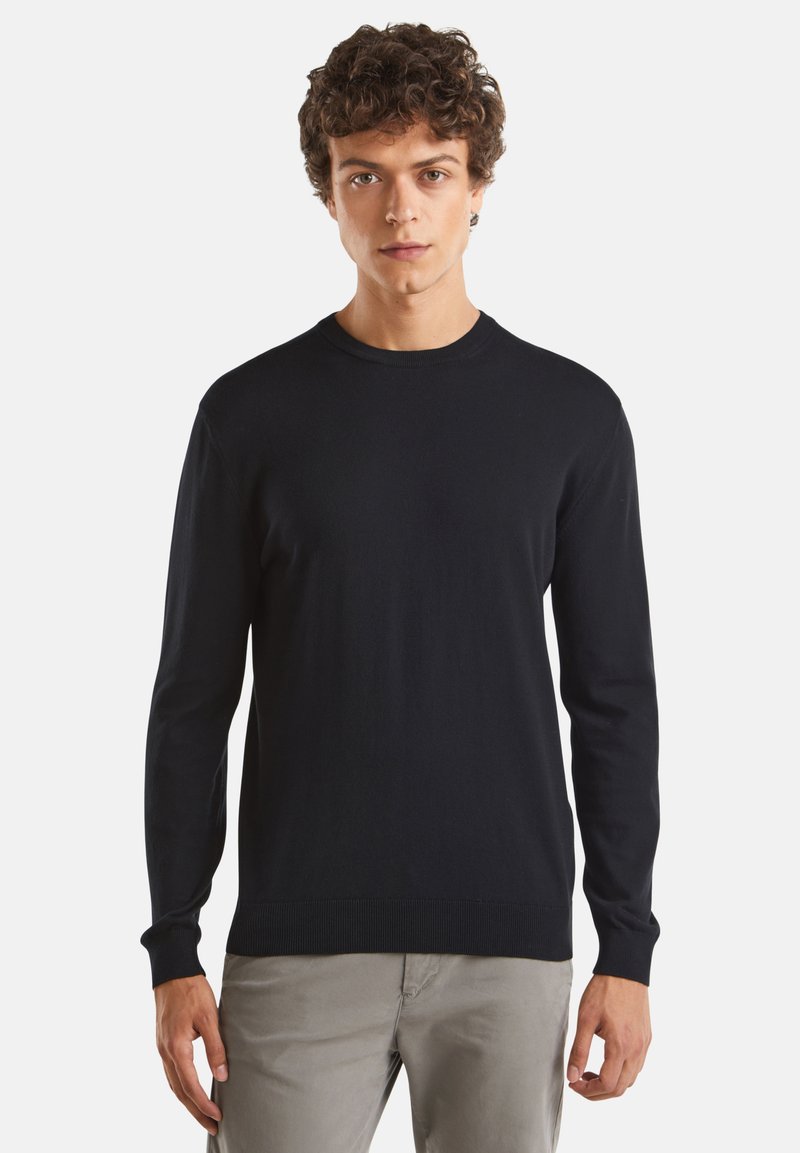 Černý pletený svetr s dlouhými rukávy a kulatým výstřihem. Má hladkou texturu a žebrované manžety a spodní lem. Nosený přes světlé kalhoty.