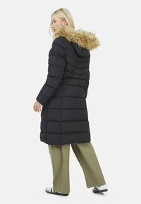 Doudoune noire avec des coutures horizontales, capuche en fourrure, et manches longues. Assortie avec un pantalon large vert olive et des baskets noires.
