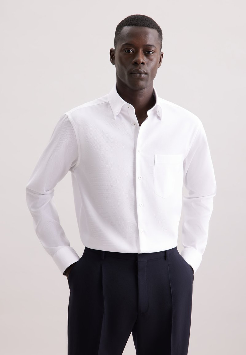 Uomo con capelli corti che indossa una camicia bianca a maniche lunghe e pantaloni scuri, in piedi con le mani nelle tasche su uno sfondo semplice.