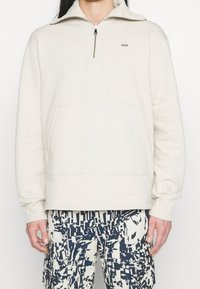 Světlé béžové mikina na zip s vysokým límcem, přední kapsou a vyšitým logem W.W. Měkká, texturovaná látka.