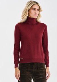 Bordeauxroter Rollkragenpullover aus weichem Strickstoff, mit langen Ärmeln und einem gerippten Saum, versehen mit einem kleinen Markenzeichen am unteren Rand.