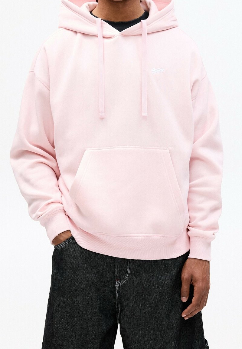 Personne portant un sweat à capuche rose clair avec poche avant et pantalon noir, les mains partiellement dans les poches, sur un fond uni.