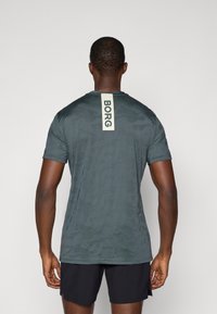 Σκούρο πράσινο αθλητικό μπλουζάκι με καμουφλάζ σχέδιο, κοντά μανίκια και κάθετη λευκή ετικέτα λογότυπου πίσω. Το υλικό φαίνεται ελαφρύ.